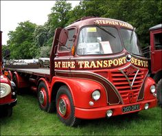 an old red transport truck is parked on the grass in front of other antique trucks