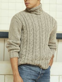 a man wearing a turtle neck sweater standing in front of a white tiled wall with his hands in his pockets