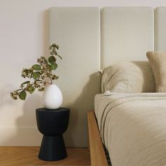 a bed with a white headboard next to a plant on a black stand in front of it