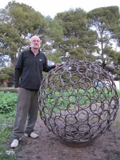 a man standing next to a large metal object in a field with trees behind him