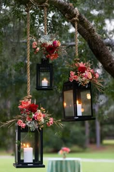 three hanging lanterns with flowers and candles are hung from a tree branch in front of a table