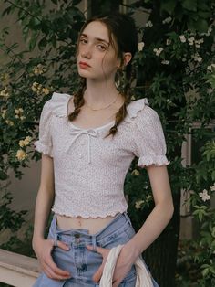 a woman with braids standing in front of some bushes and flowers wearing a white top