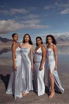 three women in white dresses standing next to each other on the beach with mountains in the background