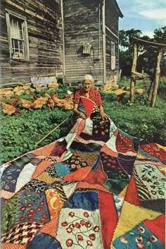 an old woman sitting on the grass next to a house with many different quilts