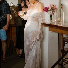 a woman in a white dress standing next to a wall holding a cup and looking at her phone