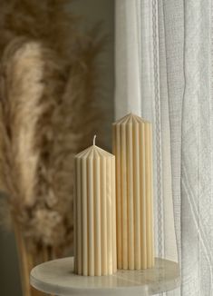 two white candles sitting on top of a table next to a window with sheer curtains