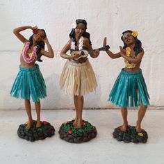 three figurines are standing next to each other on a white surface, one is holding a guitar and the other two are wearing grass skirts