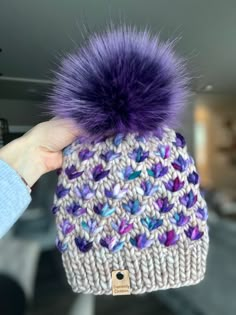 a hand holding up a knitted hat with purple pom - pom