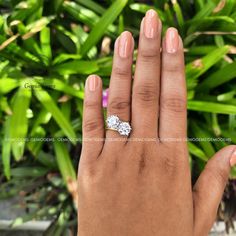 a woman's hand with a diamond ring on top of her finger and green plants in the background