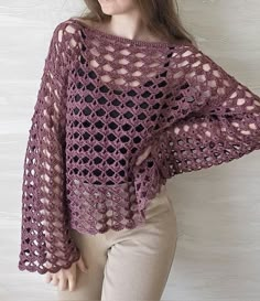 a woman wearing a purple crocheted sweater and tan pants standing in front of a white wall