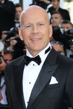 a bald man in a tuxedo and bow tie smiles as he poses for photographers