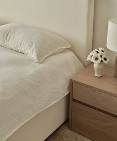 a white vase with flowers sitting on top of a bed next to a night stand