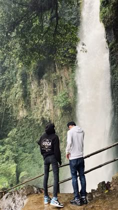 two people standing at the edge of a cliff looking at a waterfall in the distance