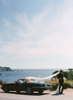 a bride and groom standing next to an old black car on the side of the road