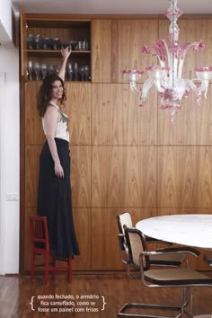 a woman standing in front of a dining room table with chairs and a chandelier