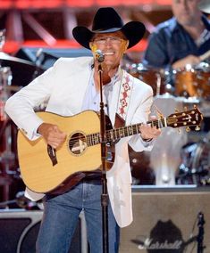 a man wearing a cowboy hat and holding a guitar in front of a microphone on stage