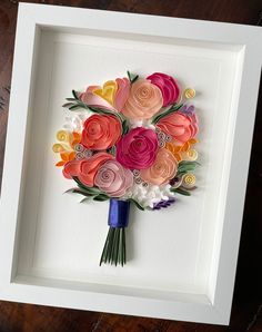 a paper flower bouquet in a white frame on a wooden table next to a wall