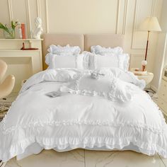 a white bed with ruffles and pillows in a room next to a chair