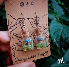 a pair of tiny glass jars filled with moss and mushrooms are held in front of a card that says ghost's little shop