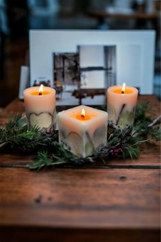 three candles sitting on top of a wooden table next to greenery and a white box