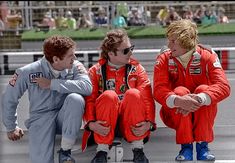 three men in red racing suits sitting on the ground with their feet up and hands clasped