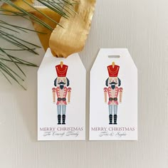 two christmas gift tags sitting on top of a white table next to a pine branch