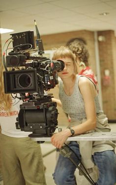 a woman is sitting down with a camera in front of her and another person standing behind her