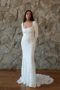 a woman standing in front of a stone wall wearing a white wedding dress with long sleeves