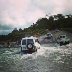 several vehicles are crossing a river in front of a hill and people on the other side