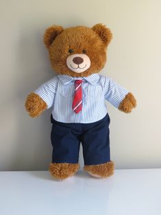 a brown teddy bear wearing a blue shirt and red tie with dark blue pants on a white table