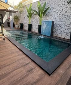 an outdoor swimming pool with water feature and plants on the side wall, surrounded by wood flooring