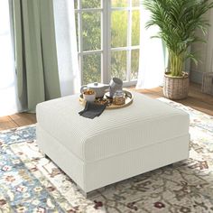 a white ottoman sitting on top of a rug in front of a window with potted plants