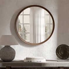 a round mirror sitting on top of a wooden table next to a lamp and book