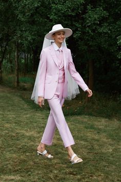 a woman in a pink suit and white hat walking on grass with trees behind her