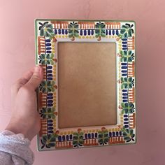 a person holding up a small photo frame with flowers on the front and sides, against a pink wall