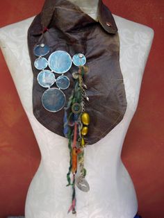 This cool bib collection is mostly recycled! The leather is remnants from a furniture maker, the blue capiz circles are repurposed from a candle holder and the brass hardware is from vintage jewelry. I cinched the leather creases with thick red thread and connected the shells with screw head anchors. The colorful yarn was hand spun suspending cast concrete charm.Â  Finished with gold top stitching Measures 10" long,(16" with fringe) 8" wide Neck measures 13" around Blue Bohemian Bib Necklace, Brass Necklace Bib, Unique Handmade Blue Bib Necklace, Bohemian Shell-shaped Brass Jewelry, Bohemian Blue Shell-shaped Jewelry, Recycle Bin, Cast Concrete