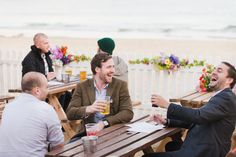 three men sitting at wooden tables talking and drinking drinks on the beach with flowers in the background