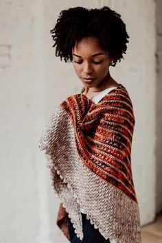 a woman standing in front of a white brick wall wearing a shawl and jeans