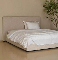 a bed with white sheets and pillows next to a potted plant on the floor