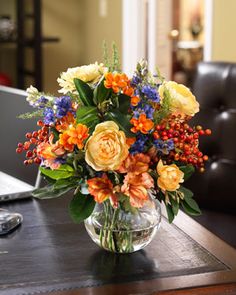 a glass vase filled with flowers on top of a wooden table next to a laptop computer