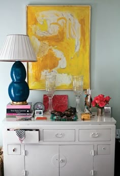 a white dresser topped with lots of drawers next to a lamp and painting on the wall