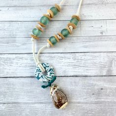 Beach-themed necklace that is ready to jump in your suitcase for that beach vacation! A 2-Inch seashell from an Indian Ocean Mix of shells hangs from a fabric-wrapped wooden hoop with a little bow made of vintage sari silk. This seashell and hoop pendant measuring 4-Inches sits center stage on this beach-inspired pendant necklace complimented with sage sea glass beads and wooden saucer spacer beads strung on 1/8-inch wide rope cord with brass end caps and lobster claw closure. This calming palet Unique Shell Necklace For Beach, Green Strand Necklace For Beach, Green Strand Necklaces For Beach, Handmade Ocean Color Necklaces For Beach, Ocean-inspired Green Necklaces For Beach, Green Ocean-inspired Necklaces For The Beach, Green Ocean-inspired Necklaces For Beach, Unique Beaded Necklace For Beach, Unique Adjustable Necklaces For Beach