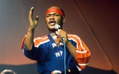 a man standing in front of a microphone with his hand up to the air and wearing an orange headband