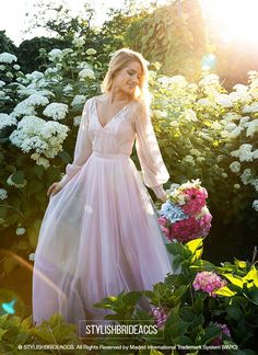 a woman in a pink dress standing next to some white flowers and greenery with the sun shining on her face