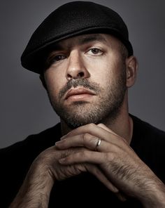 a man wearing a black hat with his hand on his chin looking at the camera