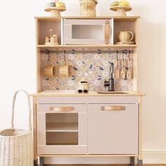 a wooden play kitchen with white cabinets and beige cupboards on the wall next to a wicker basket