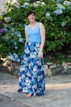 a woman wearing a blue top and floral print skirt standing in front of some flowers