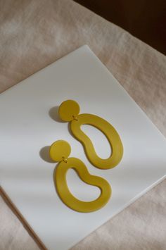 a pair of yellow earrings sitting on top of a white square piece of paper next to a beige cloth