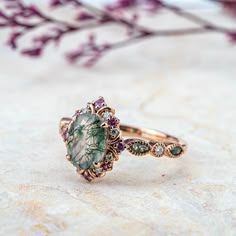 This vintage-inspired ring exudes timeless elegance with a natural oval-cut moss agate at its center, symbolizing the beauty of nature's intricate patterns. Surrounding the moss agate are round-cut moissanite stones and delicate amethysts, adding a touch of regal charm and vibrant color contrast. The ornate band is a work of art, merging classic and nature-inspired elements. Its unique combination of stones evokes a sense of serenity, growth, and mystery. Perfect for those who appreciate one-of-a-kind craftsmanship and the quiet elegance of nature. 𝗗𝗘𝗧𝗔𝗜𝗟𝗦𝗠𝗔𝗜𝗡 𝗦𝗧𝗢𝗡𝗘𝗚𝗲𝗺𝘀𝘁𝗼𝗻𝗲: Natural Moss Agate𝗖𝗼𝗹𝗼𝗿: Green𝗦𝗵𝗮𝗽𝗲: Oval 6.0*8.0mm𝗖𝘂𝘁: Full𝗠𝗲𝗮𝘀𝘂𝗿𝗲𝗺𝗲𝗻𝘁: 1.5 ct approx𝗤𝘂𝗮𝗹𝗶𝘁𝘆: VS clarity gradeThe color and texture of each natural moss agate gem Vintage Amethyst Ring Gold, Luxury Green Oval Moonstone Ring, Luxury Agate Gemstone Rings, Luxury Moss Agate Gemstone Rings, Moss Agate Rings, Regal Style, Agate Rings, Amethyst Wedding, Nature Inspired Engagement Ring