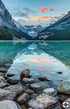 a lake surrounded by mountains with rocks in the foreground and snow capped peaks in the background